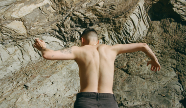 A shirtless person with tattoos on their arms climbs a textured rock face, facing the rock with arms stretched wide for grip.