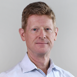 A man with short reddish-brown hair wearing a light blue collared shirt, posed against a plain light background.