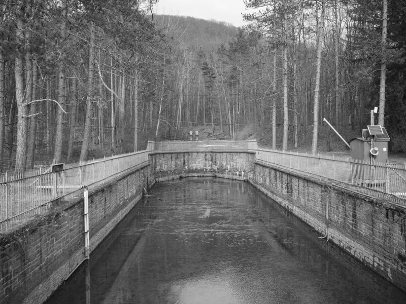A black-and-white photo shows a rectangular canal bordered by stone walls and metal railings, surrounded by leafless trees. A small control building with solar panels stands on the right side.