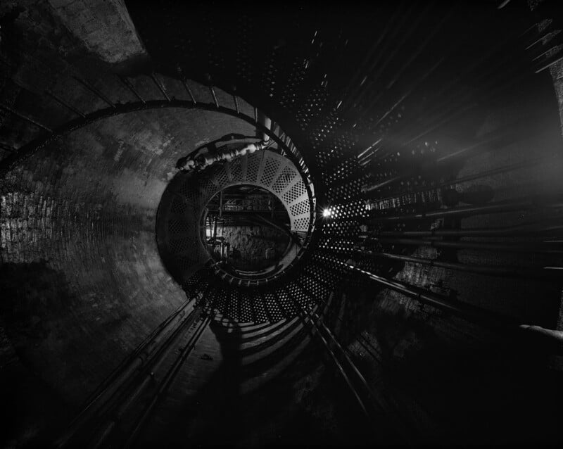 A spiral metal staircase descends into a dark, cylindrical industrial space, illuminated by dramatic lighting that casts strong shadows and reveals a network of pipes along the curved concrete walls.