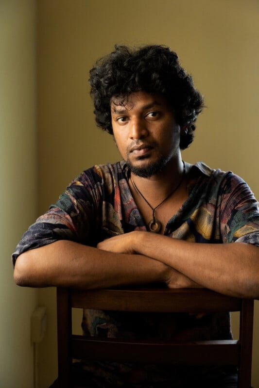 A man with curly hair and a beard sits on a chair facing backward, resting his arms on the chair’s back. He wears a patterned shirt and looks at the camera with a neutral expression against a beige background.