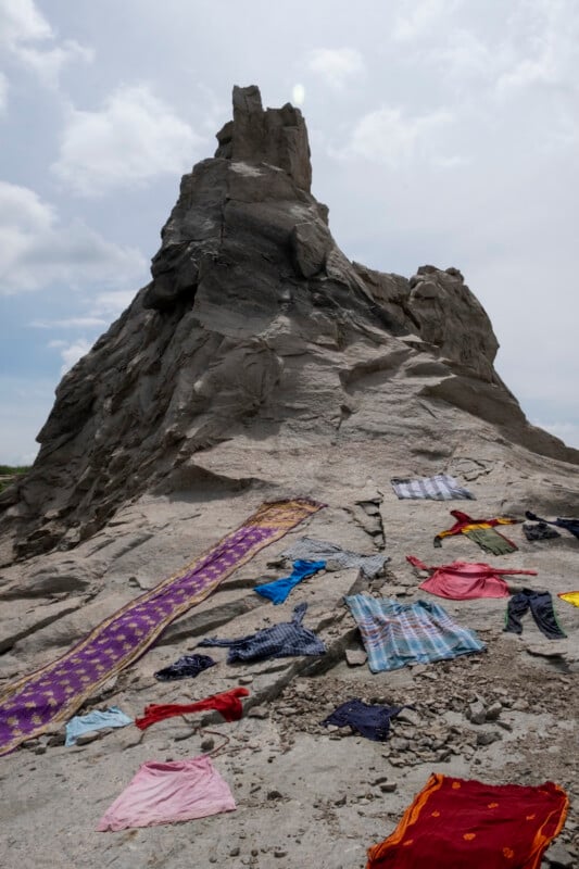 Clothes and colorful fabrics are spread out on rocky ground in front of a tall, jagged rock formation under a cloudy sky, possibly drying or displayed.