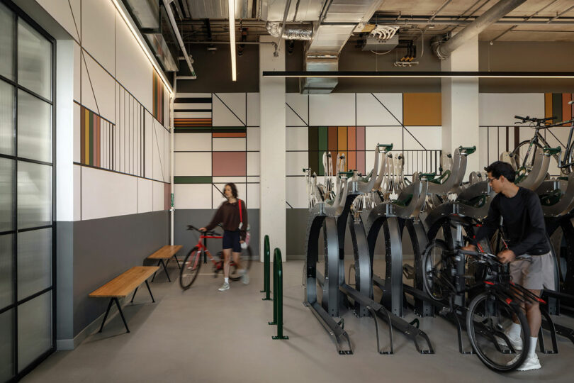 Two people with bicycles stand in a modern indoor bike storage room designed by Morris Adjmi Architects, featuring geometric wall art, benches, and bike racks.