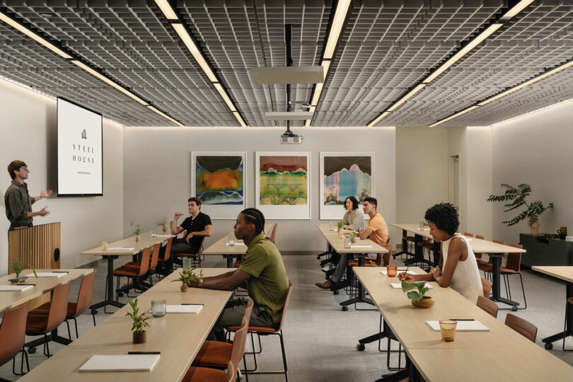 A presenter stands at the front of a modern conference room designed by Morris Adjmi Architects, speaking to six seated attendees. The room features tables, plants, wall art, and a screen displaying "Steel House.