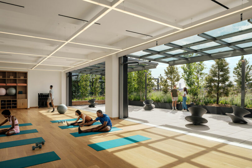 Modern fitness studio by Morris Adjmi Architects with large windows, people stretching on yoga mats indoors, and two outside on a terrace with greenery and sculptural seats. Natural light fills the space.