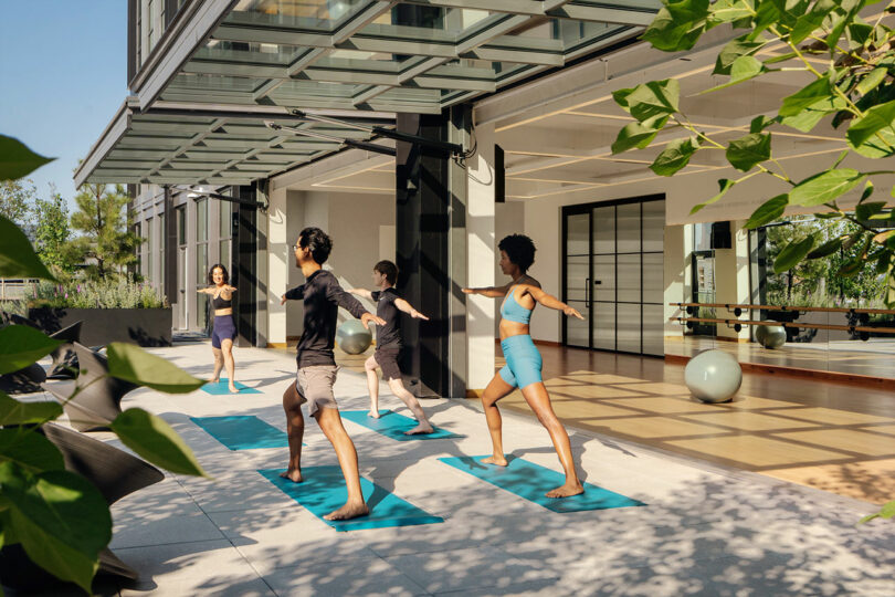 Four people practice yoga on blue mats outside a modern building designed by Morris Adjmi Architects, with large glass doors. Indoor exercise balls and surrounding greenery add to the tranquil scene.