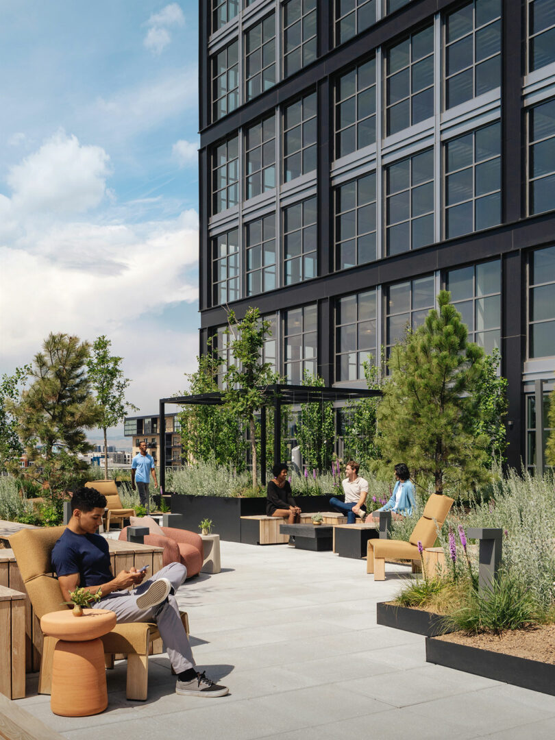 People sit and relax on modern outdoor furniture surrounded by greenery on a rooftop terrace next to a tall, glass-paneled building designed by Morris Adjmi Architects.