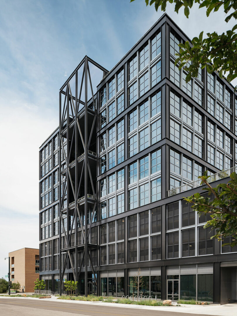 Modern multi-story office building by Morris Adjmi Architects, with a black steel and glass facade, external structural supports, and large windows, surrounded by a few trees under a clear sky.