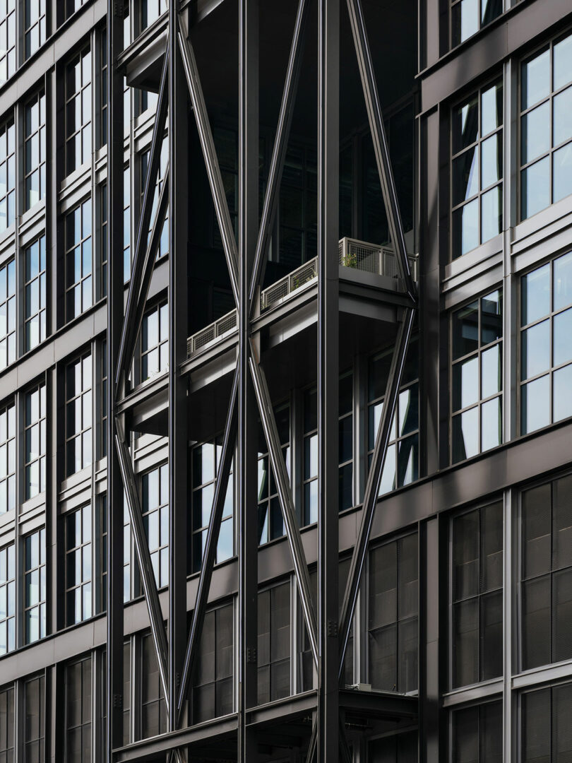 Modern building facade by Morris Adjmi Architects featuring glass windows, black metal beams in an X-shaped pattern, and a balcony with a small railing and greenery.