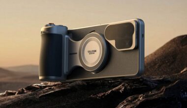 A smartphone with a large camera and ergonomic grip attachment rests on a rocky surface with a desert landscape in the background during sunset.