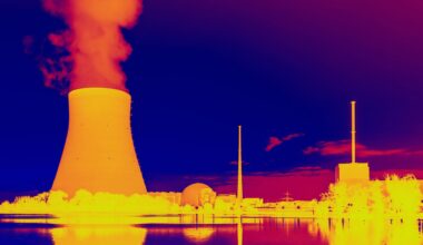 This infrared image shows a glowing orange nuclear power plant releasing hot yellow smoke near various outbuildings with blue water and sky above and below
