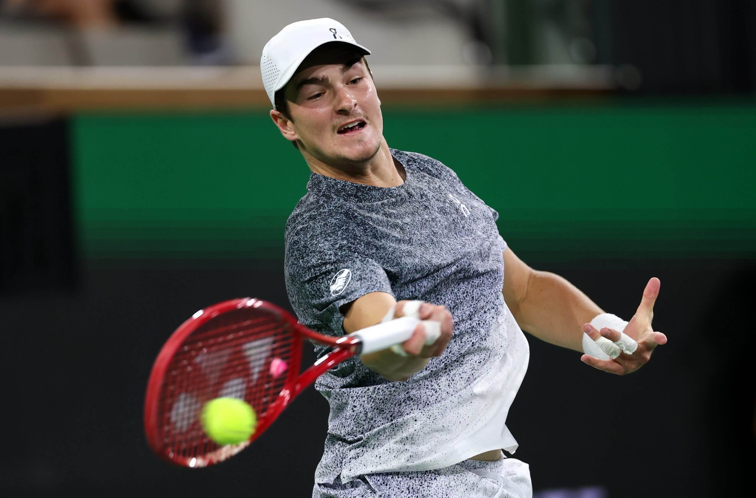 João Fonseca hits a forehand with a red tennis racket, wearing a grey-and-white outfit and a white hat.