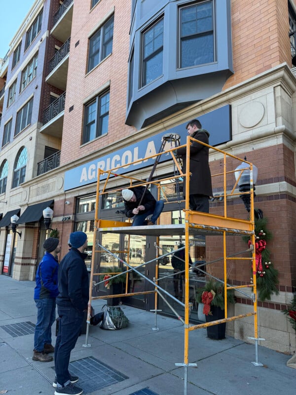 Four people work with scaffolding outside a brick building labeled "AGRICOLA." Two are on the scaffold adjusting a sign, while two others stand on the sidewalk observing. Holiday wreaths decorate the entrance.