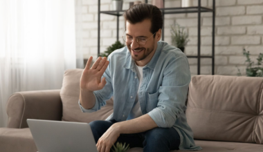 A man sitting on a couch, using a laptop for V A TelePulmonology, and waving with one hand, smiling at the camera.