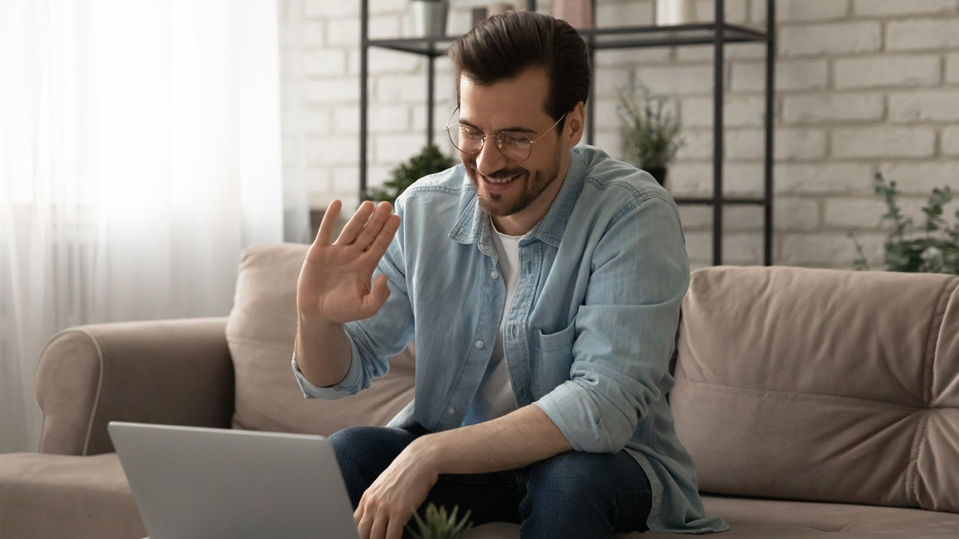 A man sitting on a couch, using a laptop for V A TelePulmonology, and waving with one hand, smiling at the camera.