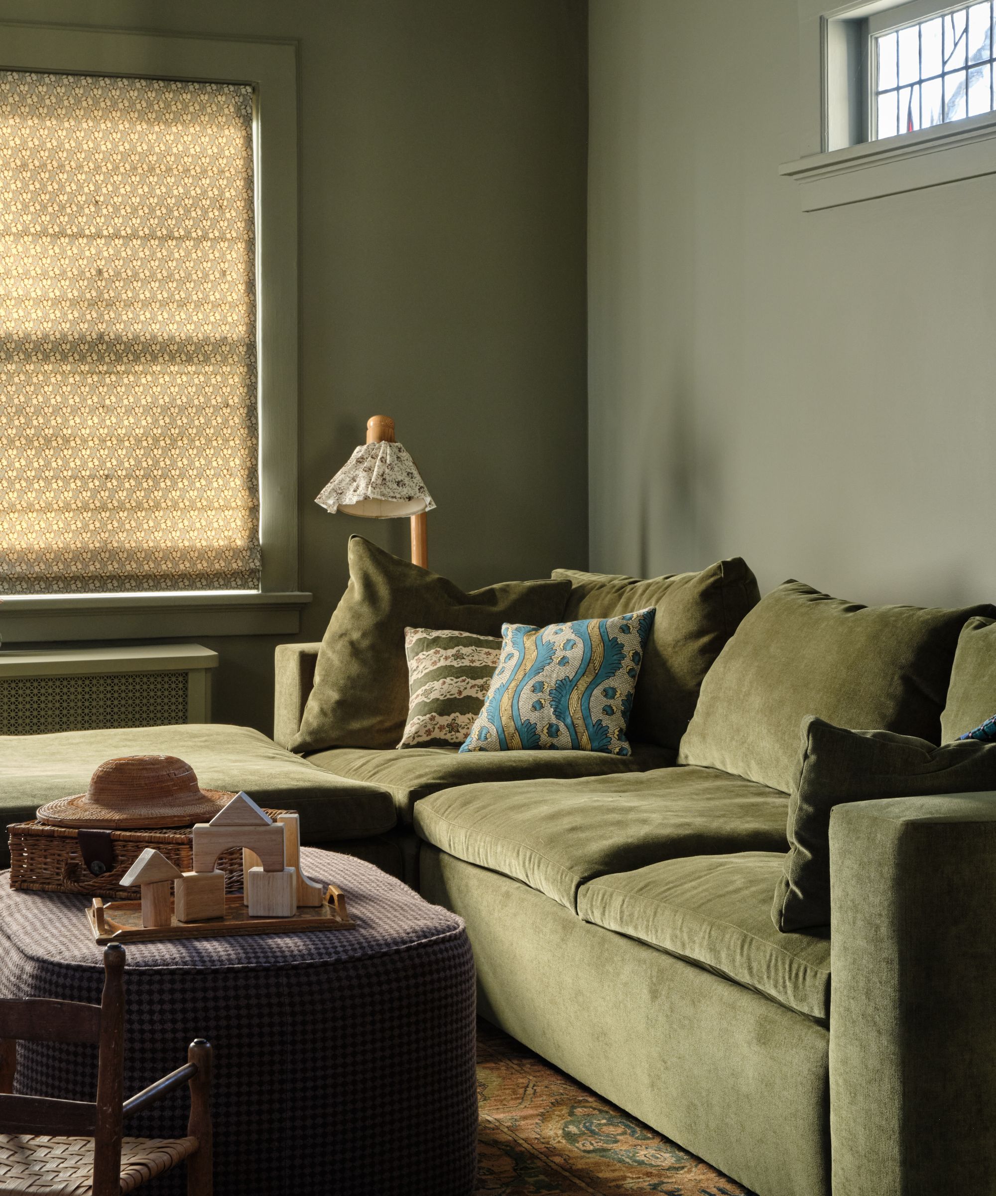 Lounge with dark green sage walls, dark green velvet sofa, roman blind on window, gingham and ottoman with toys on top