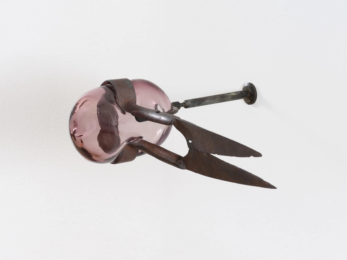Sculpture of a pink glass orb held by rusty metal clamps mounted on a wall.