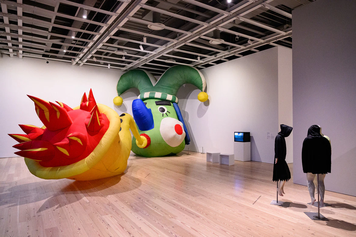 Art gallery with large inflatable sculptures and two mannequins in black clothing on a wooden floor.