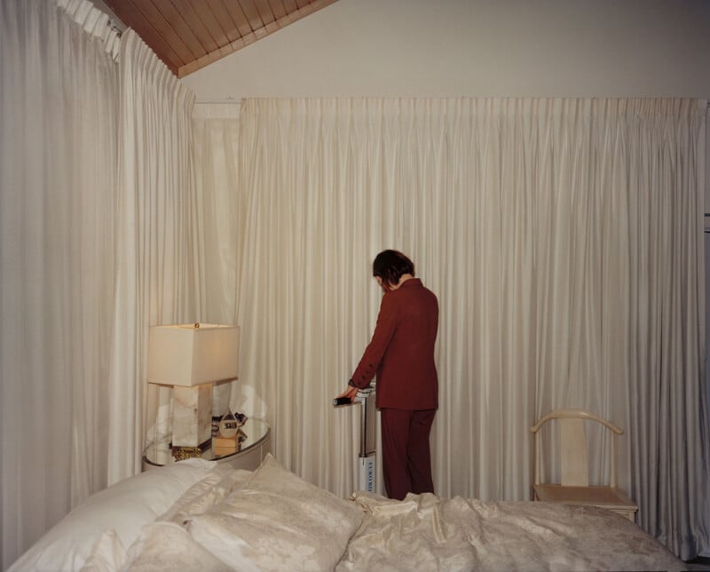 A person in a red suit stands facing drawn cream-colored curtains in a softly lit bedroom with a bed, a lamp, and a wooden chair visible. The person’s face is not shown.