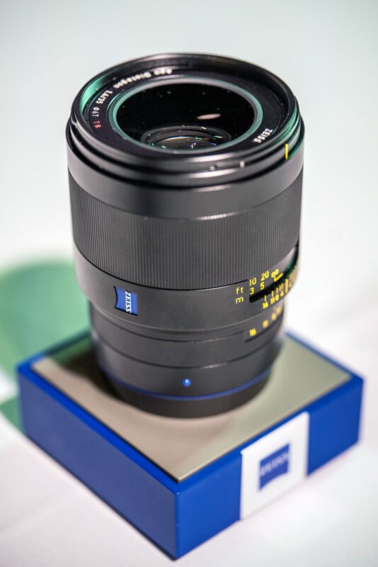 A Zeiss camera lens is displayed upright on a blue and white box, with focus and distance markings visible on the lens barrel against a softly blurred background.