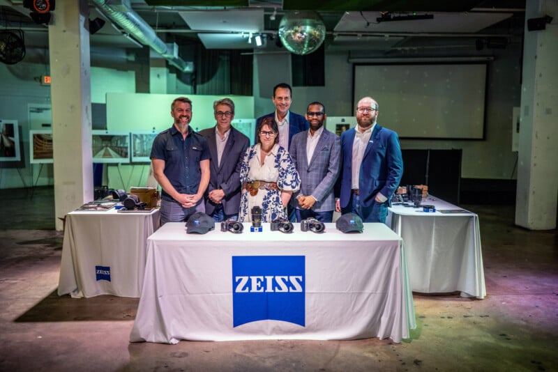 Six people stand behind a Zeiss-branded table displaying cameras and equipment in a modern, industrial-style room with a disco ball overhead. The group is smiling and dressed in business or smart-casual attire.