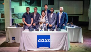 Six people stand behind a table covered with a white cloth featuring the blue ZEISS logo, displaying cameras and gear, in a modern indoor event space with exhibition panels in the background.