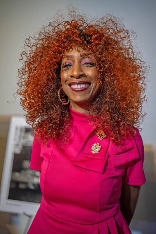 A woman with vibrant curly red hair and a bright smile wears a vivid pink dress with a large bow and jeweled brooch, standing indoors with a blurred background.