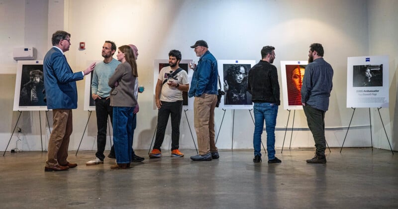 A group of people stand and converse in an art gallery, viewing portraits displayed on easels against a white wall. The atmosphere is casual and social.