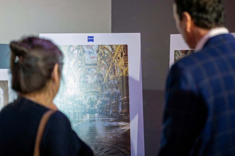 Two people stand closely together, looking at a large, framed photo of an ornate, grand interior with gilded ceilings and intricate architectural details displayed on an easel.