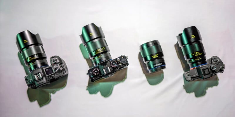 Four cameras with attached lenses and one standalone lens are arranged on a white surface, viewed from above. Each camera and lens is positioned parallel, casting soft shadows.