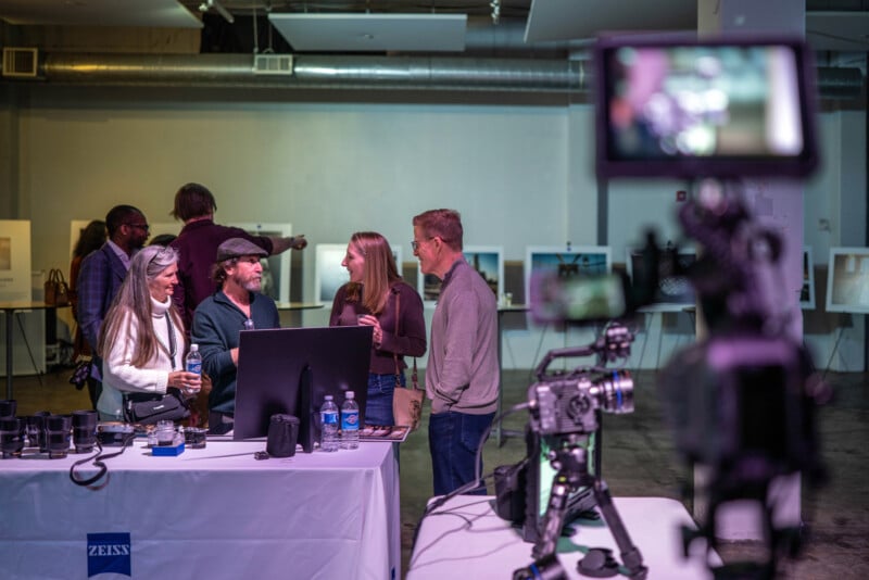 A group of people smiles and chats around a display table with cameras and lenses at an indoor event, while filming equipment and a camera in the foreground film the scene.