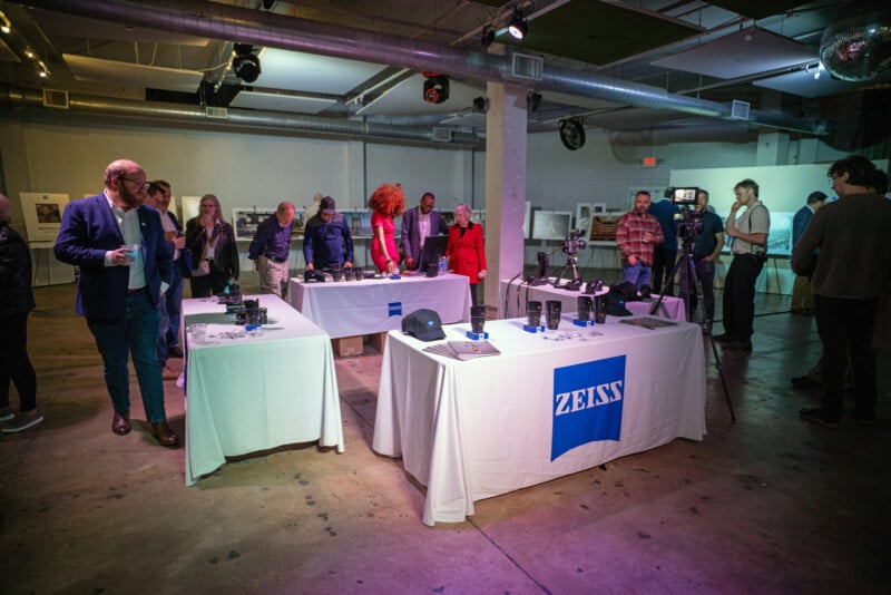 People gather around tables displaying camera equipment with ZEISS logos at an indoor event. Some attendees examine items, while others converse. The space is well-lit, industrial, and features exposed pipes.