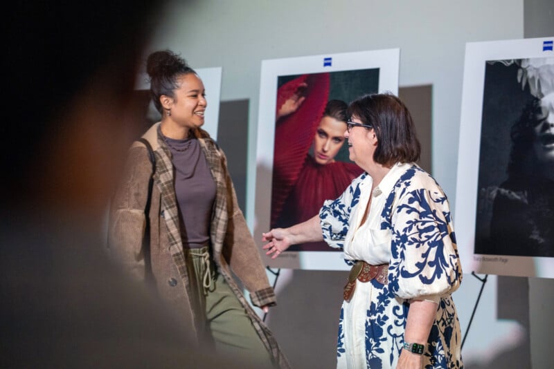 Two women smiling and interacting in front of displayed portrait photographs; one wears a brown plaid coat and green pants, the other wears glasses and a white dress with blue patterns.