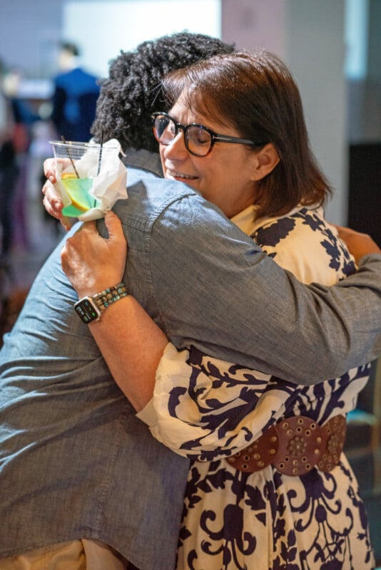 A woman in glasses and a patterned dress hugs a person in a denim shirt. The woman smiles warmly and holds a drink with a napkin in one hand. The background is softly blurred.