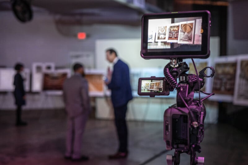 A camera on a tripod films two people in suits having a conversation in a blurred indoor setting; the camera’s display screen shows a focused view of the scene.