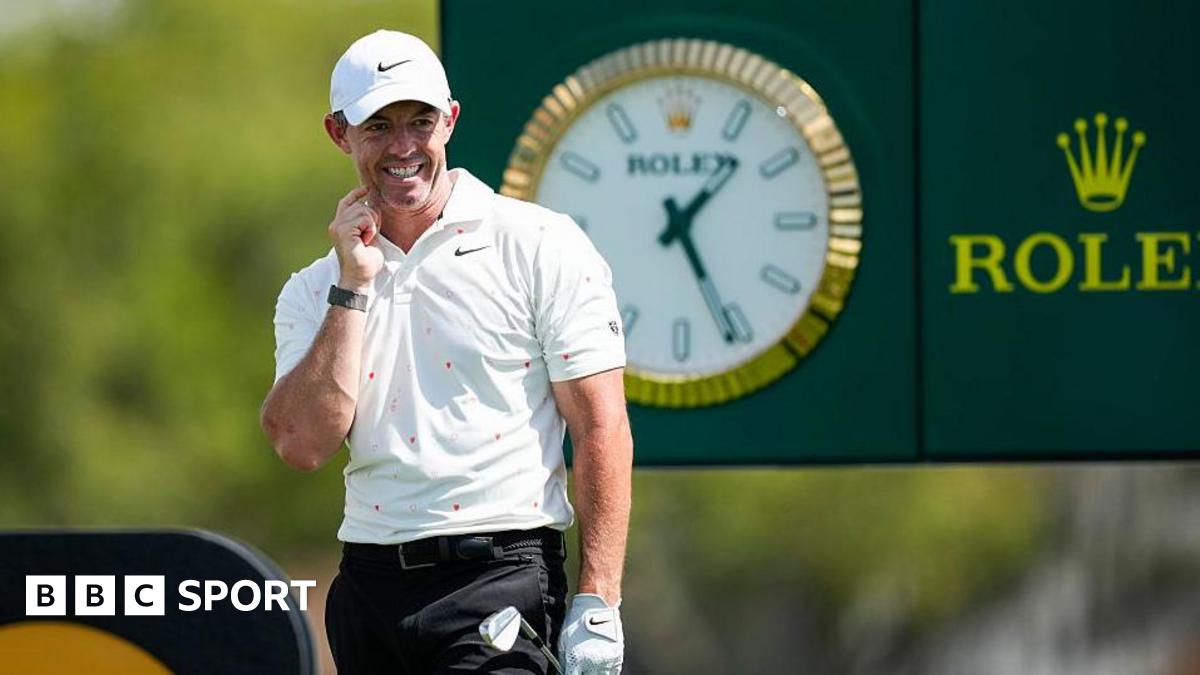 Rory McIlroy looks on after firing his tee-shot at the 14th hole of the Arnold Palmer Invitational. He is wearing a white cap, white Nike polo shirt and black trousers. He is holding his club in his gloved left hand. A large Rolex clock and advertising board can be seen behind him.