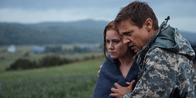 Amy Adams and Jeremy Renner as Louise and Ian in 'Arrival' stand outside in a grass field holding each other. Louise is wrapped in a blanket and her hair is wet.
