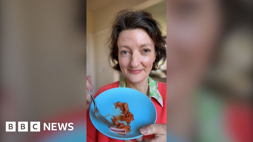 A woman with brown hair holds up a blue plate holding kimchi. She is wearing a dark pink jumper and a green-collared shirt.