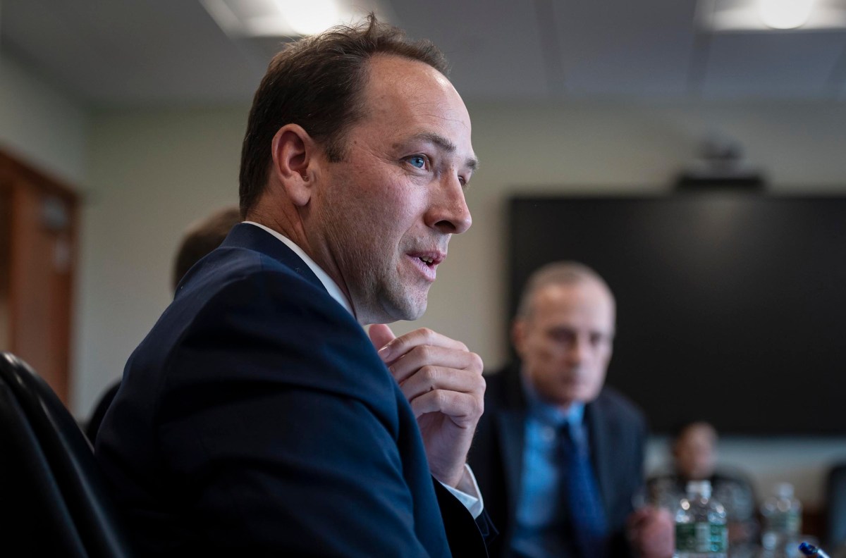 A man in a suit is speaking in a meeting room while another man, also in a suit, is seated in the background.