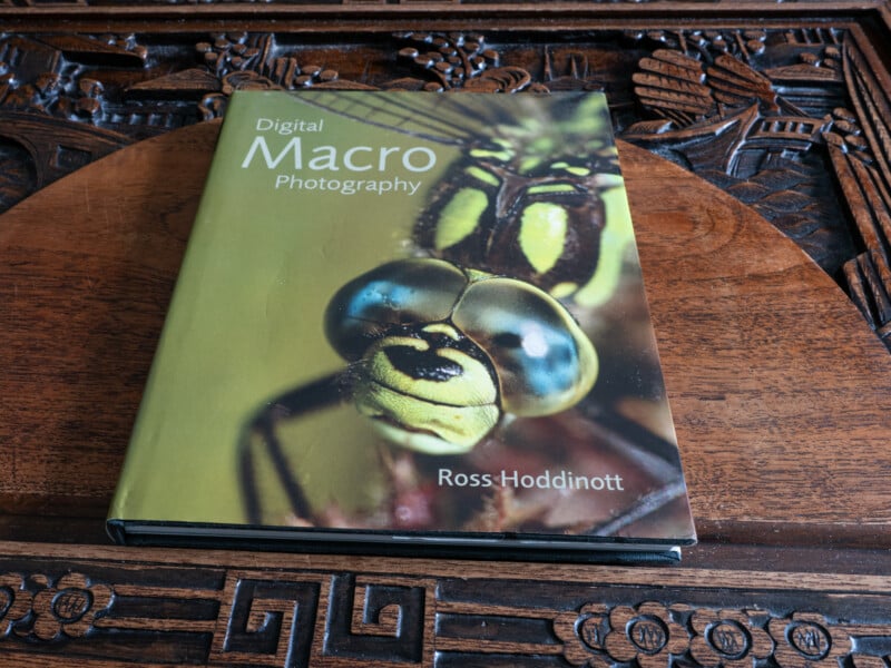 A book titled "Digital Macro Photography" by Ross Hoddinott, featuring a close-up image of a dragonfly on the cover, is placed on a carved wooden surface.