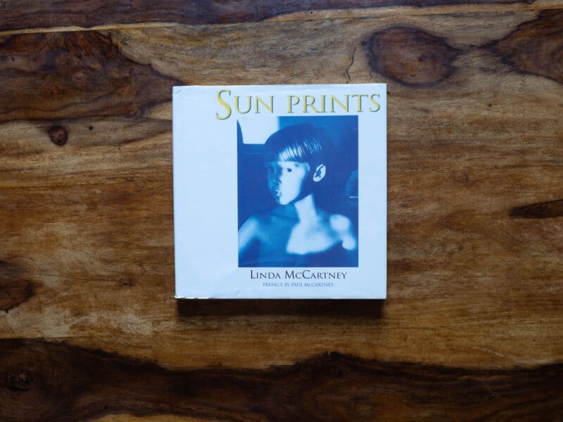 A book titled "Sun Prints" by Linda McCartney lies on a wooden surface. The cover features a blue-tinted photo of a young boy and includes a preface by Paul McCartney.