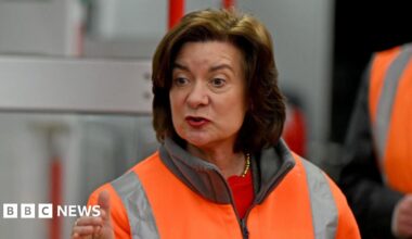 Eluned Morgan wearing a hi-viz jacket, guestering with one hand while she speaks. She is stood in front of a train which is out of focus.