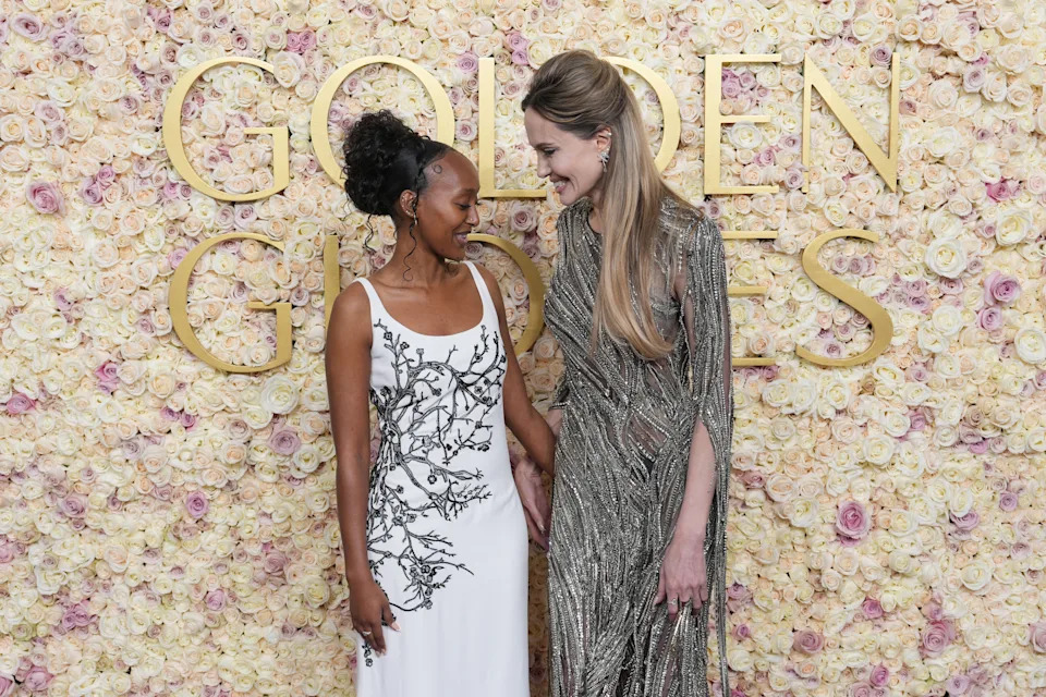 Two women stand in front of a floral backdrop. One wears a white dress with black detailing, the other a shimmering metallic gown