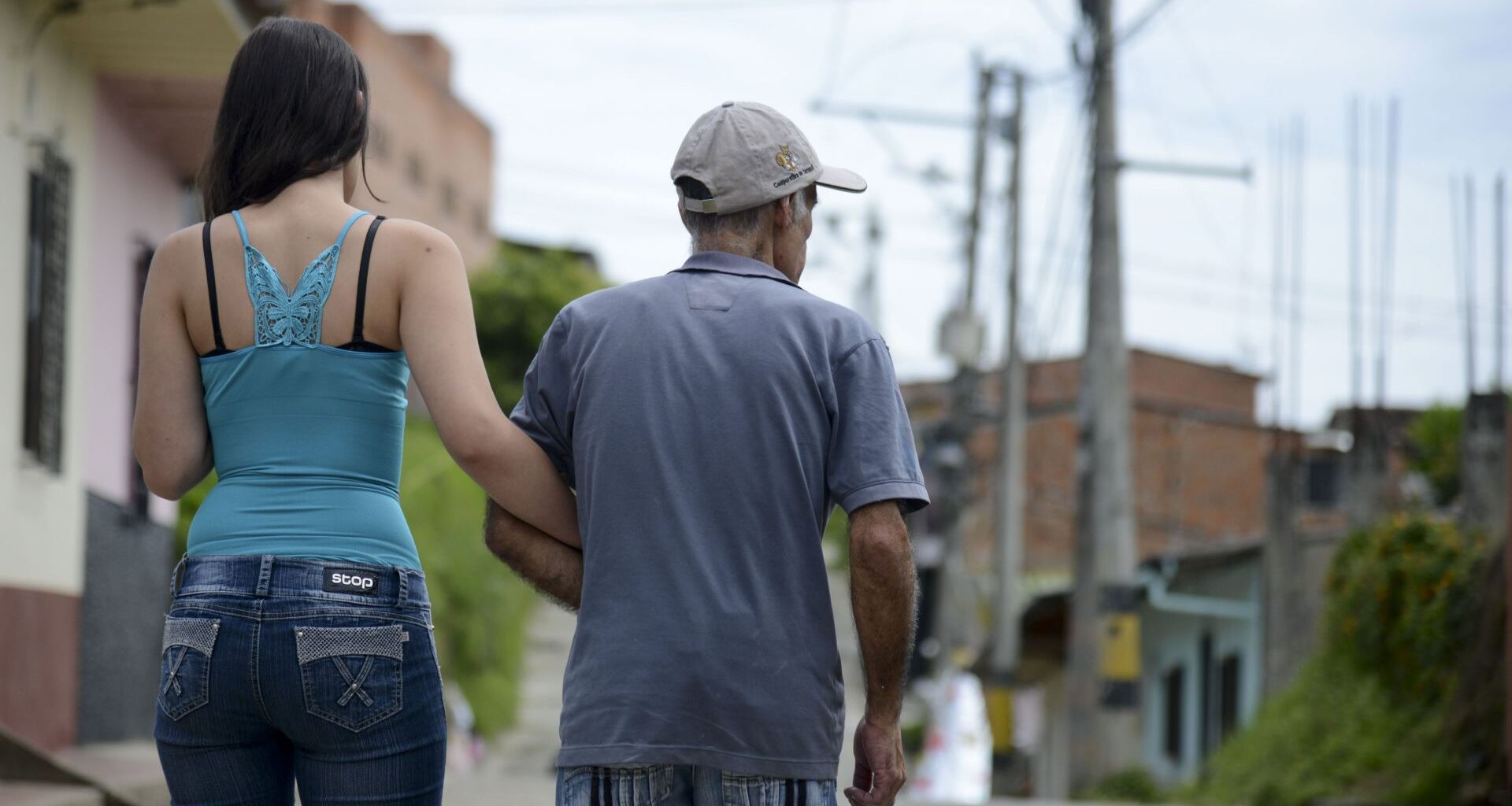 A woman wearing a blue tank top and jeans with long dark hair holds the arm of an older tan man wearing a gray shirt and a baseball cap as they walk down the street, their backs to the camera