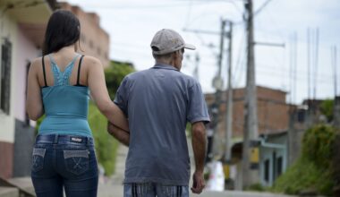 A woman wearing a blue tank top and jeans with long dark hair holds the arm of an older tan man wearing a gray shirt and a baseball cap as they walk down the street, their backs to the camera