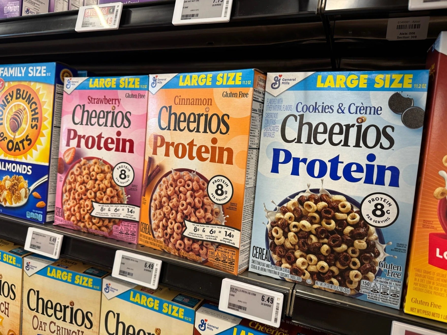 Three varieties of large size Cheerios Protein cereal boxes—Strawberry, Cinnamon, and Cookies & Creme—are displayed on a grocery store shelf with price tags below.