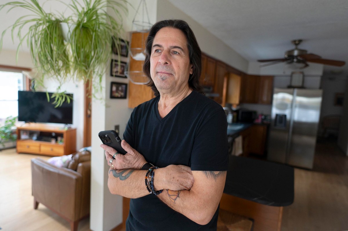 A man with long dark hair, wearing a black shirt, stands in a kitchen holding a smartphone, with his arms crossed. Houseplants and furniture are visible in the background.