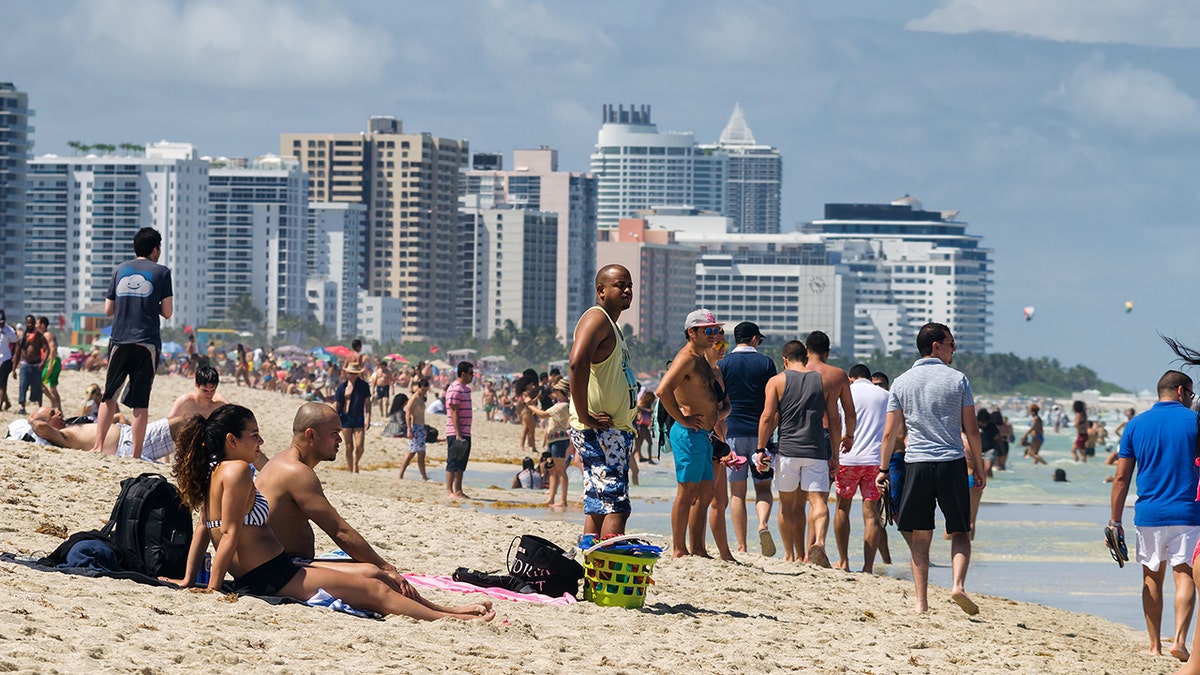 Crowded Miami Beach shoreline with sunbathers and swimmers in front of high-rise hotels and condos.