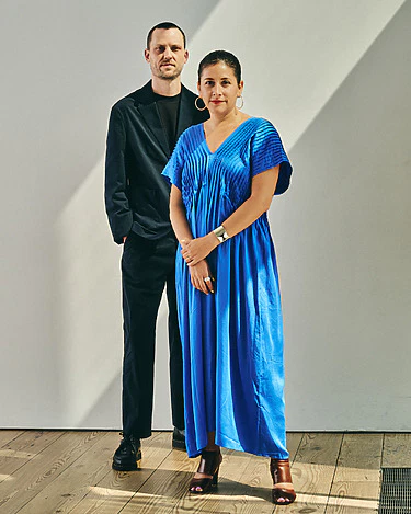 Man in black suit and woman in a blue dress standing on a wooden floor with a neutral background.