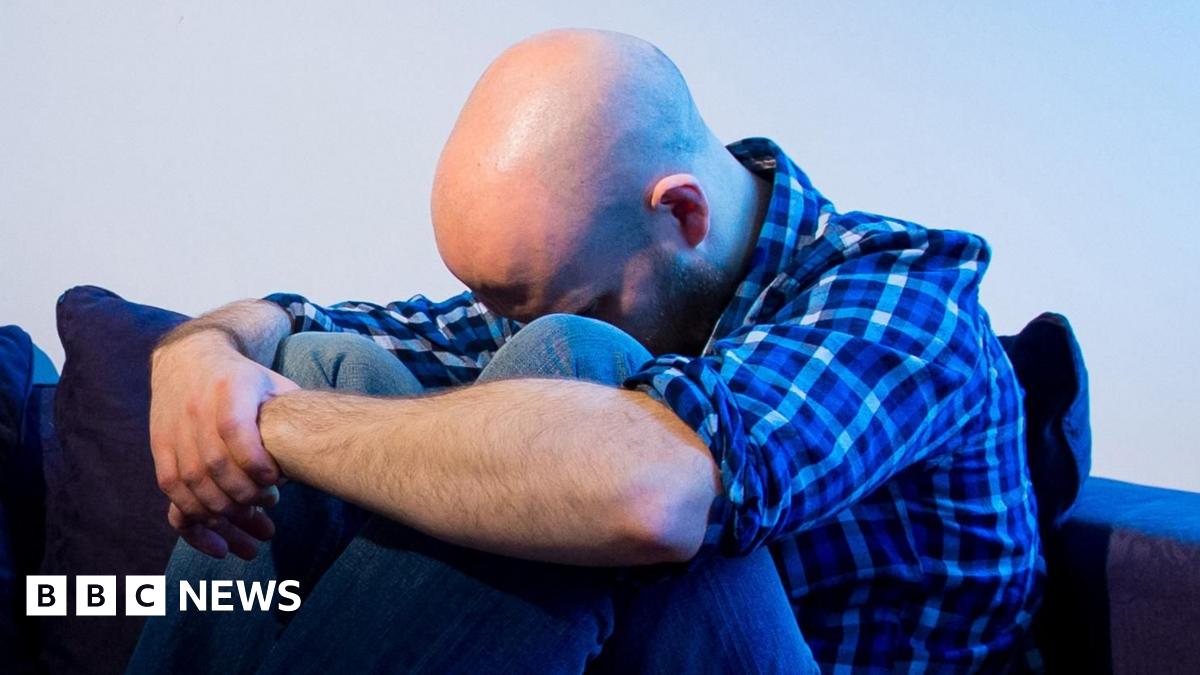 A man is hunched over on a sofa with his head between his knees and his arms locked around them.